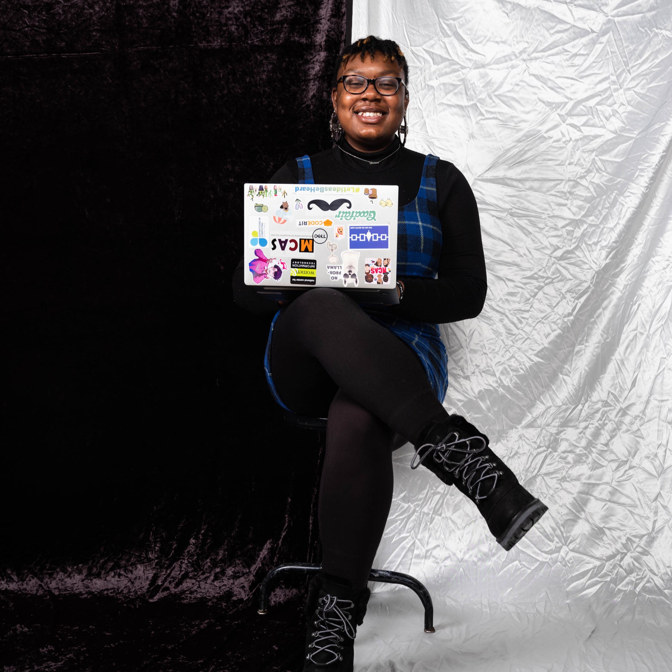 An image of me, Jabrecia, seated in a chair wearing a blue and black dress, black leggings, and black boots. There is a silver laptop on my lap covered in stickers.